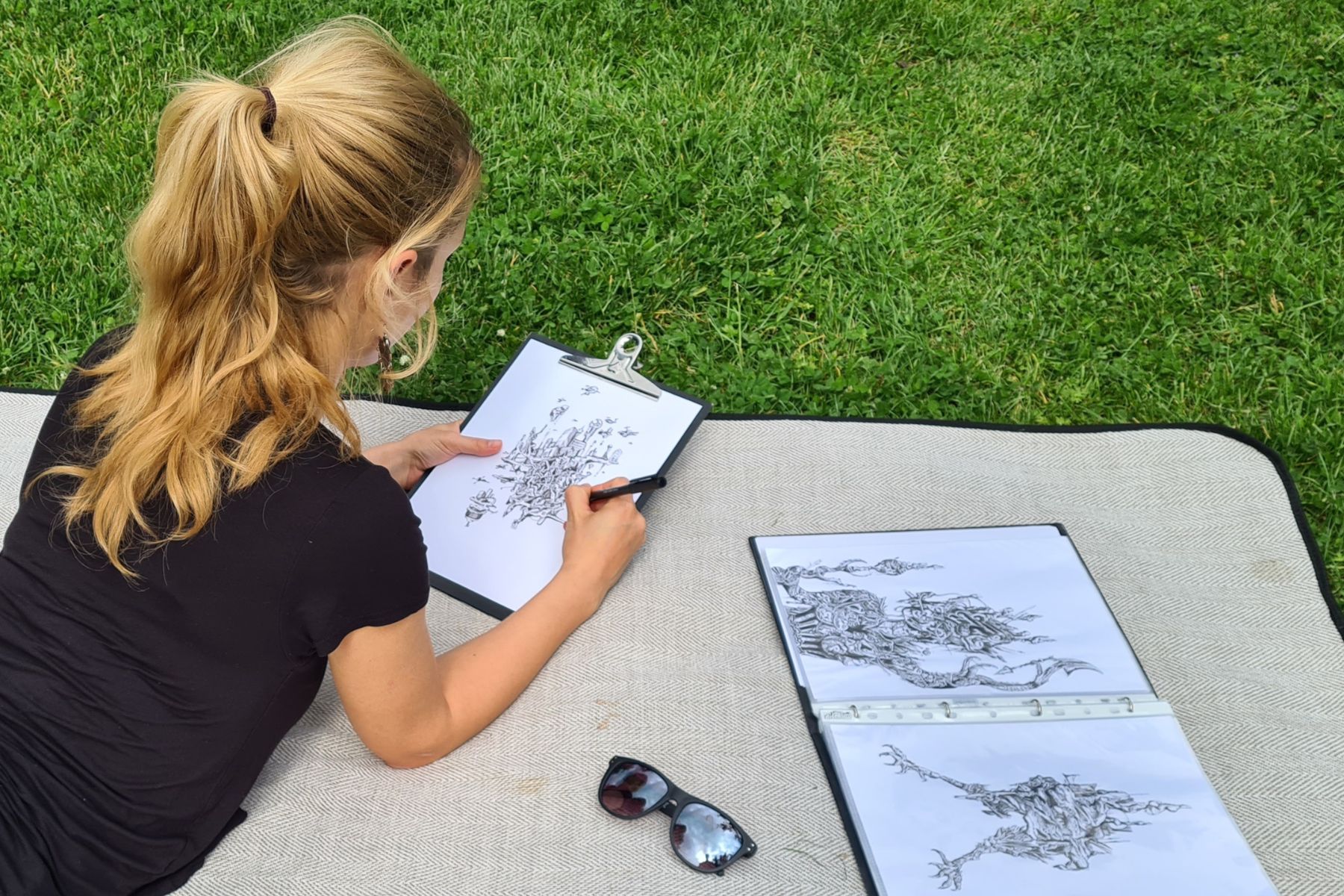 A woman lying on a blanket on the grass, deeply engrossed in drawing intricate designs on a clipboard. Next to her are additional completed sketches, showcasing detailed and imaginative artwork. Sunglasses and a pen rest nearby on the blanket. This image captures a moment of outdoor creativity and relaxation, highlighting the artistic process. It's a perfect representation for the "about artmefree" section, illustrating how inspiration can strike anywhere, turning simple doodles into intricate masterpieces.