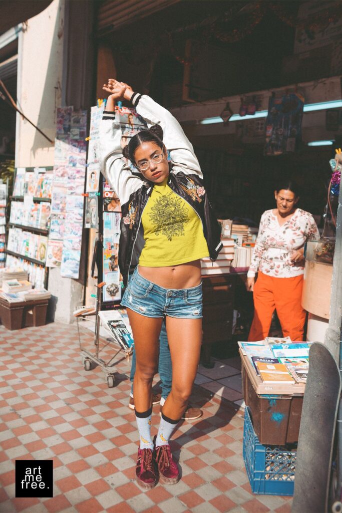 A young woman confidently stretches her arms above her head while standing in front of a bustling newsstand, capturing the essence of urban vibrancy. She exudes a carefree, stylish vibe in her black and white bomber jacket, yellow t-shirt featuring the intricate "rupture" design by artmefree, denim shorts, and burgundy sneakers paired with quirky white socks. Her hair is styled in playful buns, and she sports trendy glasses, adding to her effortlessly cool appearance. The background is lively, with an older woman curiously observing and shelves brimming with colorful magazines and books. The scene is filled with energy, embodying a mix of casual street style and creative expression. The artmefree logo in the bottom left corner signifies the artistic influence in her outfit.