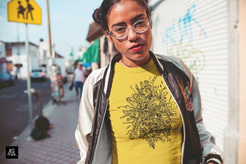 A young woman with a confident expression stands on a lively city street, creating an urban and vibrant atmosphere. She wears a black and white bomber jacket over a yellow t-shirt featuring the intricate "rupture" design by artmefree, paired with clear-framed glasses that highlight her stylish look. Her hair is styled in two neat buns, and she wears bold red lipstick that adds a pop of color to her ensemble. The detailed doodle art on her t-shirt draws attention, making it a true statement piece. The background features a busy street with people walking, a yellow pedestrian sign, and buildings adorned with graffiti, capturing the essence of city life. The artmefree logo in the bottom left corner ties the scene together, emphasizing the brand's creative influence.