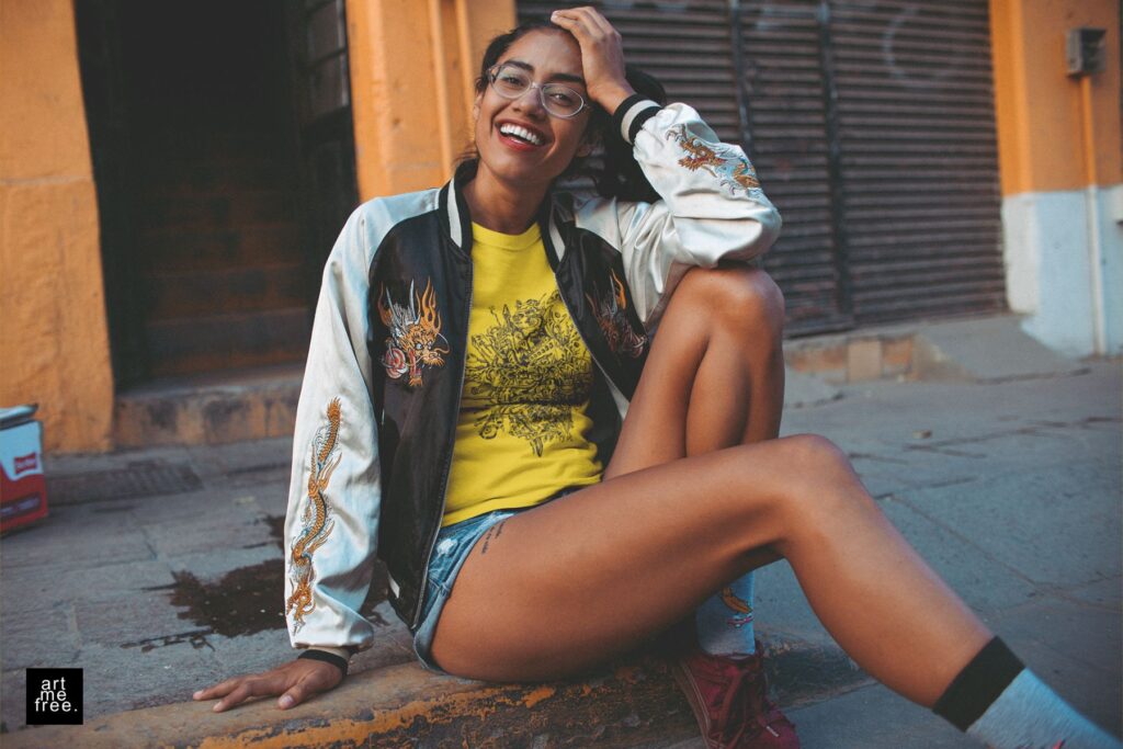 A young woman with a bright smile sits casually on a city sidewalk, exuding a relaxed and joyful vibe. She wears a black and white bomber jacket with intricate dragon embroidery over a yellow t-shirt featuring the detailed "rupture" design by artmefree. Her outfit is completed with denim shorts, burgundy sneakers, and white socks, creating a stylish and laid-back look. Her hair is styled in playful buns, and she sports clear-framed glasses that add to her trendy appearance. The background shows an urban setting with an orange building and a partially open garage door, enhancing the street-style feel of the scene. The artmefree logo in the bottom left corner subtly emphasizes the brand's creative influence.